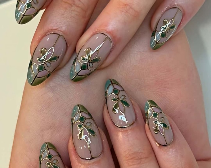 Almond-shaped nails with a nude base, featuring gold outlines and green and pink floral designs. The tips are painted in a gradient of green and blue, with gold accents. A stylish manicure.