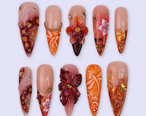 A collection of ten artificial nails with various designs. The nails feature a nude base with intricate details, including floral embellishments, gold accents, and gradient color schemes in shades of 