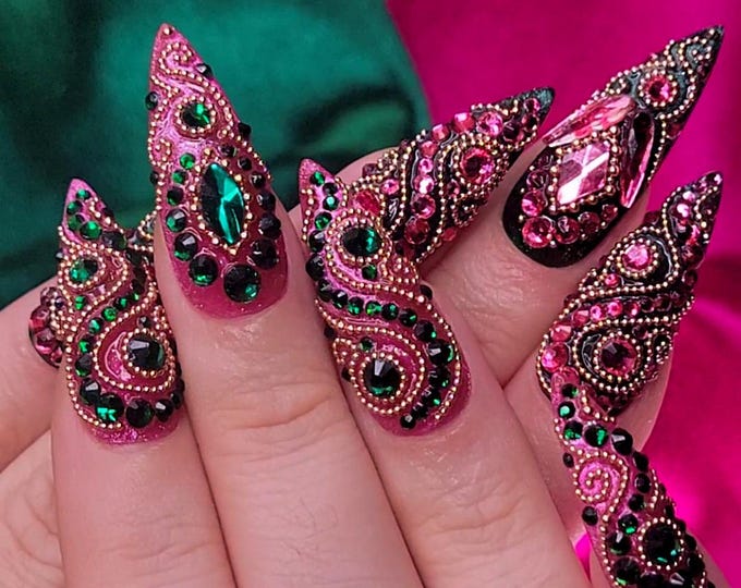 Close-up of elaborately decorated, long, pointed fingernails. The nails are painted a shimmering pink and adorned with intricate patterns of green and pink gemstones, and gold-colored embellishments. 