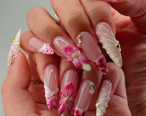 A hand displaying long, sculpted acrylic nails. The nails feature a variety of designs, including 3D pink flowers, white ruffled accents, and a pink and red dotted pattern. Embellishments include gold