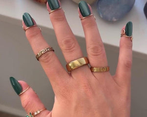 Hand with long, dark green nails and various gold rings. Rings include a thick band, a delicate butterfly ring with green accents, and several thin rings. The hand is adorned with multiple pieces of j