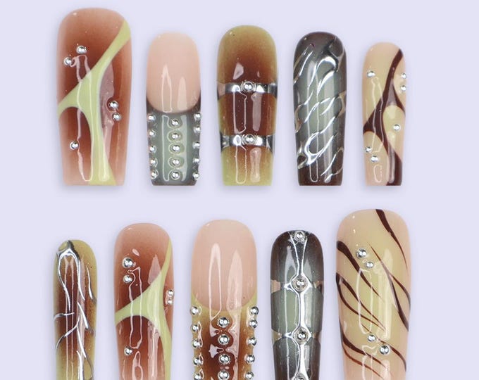 A set of ten artificial nails in various designs. The nails feature a mix of colors including beige, brown, and olive green, with decorative elements such as rhinestones and metallic accents. Some nai