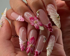 Long, sculpted artificial nails with a variety of designs. Some nails feature a pink and white floral design, while others have a French tip with a red and white pattern. Embellishments include gold b