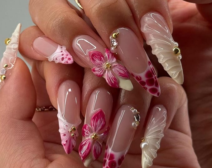 Long, sculpted artificial nails with a variety of designs. Some nails feature a pink and white floral design, while others have a French tip with a red and white pattern. Embellishments include gold b