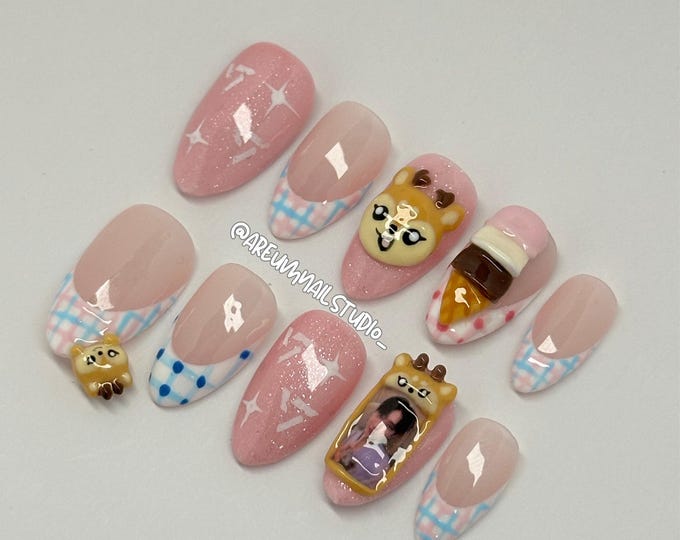 A set of artificial nails with various designs. Some nails are pink with white star patterns, others have a pink and blue plaid design. Several nails feature 3D embellishments, including a dog, an ice