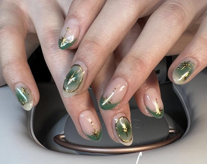 Manicured nails with a green and gold design. The nails feature a gradient green base with gold star accents and gold trim. The nails are oval-shaped and have a natural, nude base color. The word "Lux