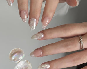 Almond-shaped nails featuring a sheer nude base with iridescent, pearlescent white tips and textured overlays. A thin silver ring with a small bow design sits on the ring finger. A small, open, irides