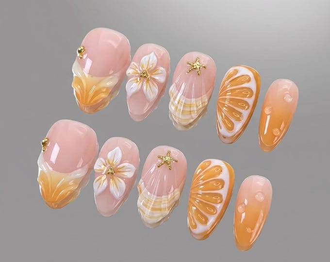 A set of ten press-on nails featuring a light pink base with orange gradient tips. Designs include white 3D flowers, gold starfish charms, small gold studs, and raised orange slice patterns with white