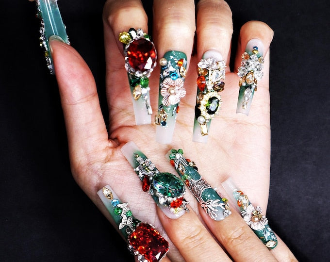 Long, square-shaped artificial nails featuring a green and white gradient base. Each nail is heavily embellished with a variety of colorful rhinestones, metallic floral charms, and faceted crystals in