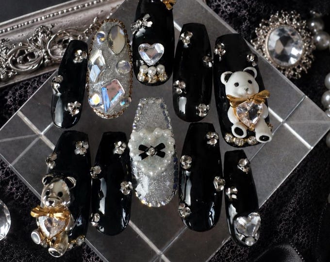 A set of ten press-on nails featuring a glossy black finish, accented with silver glitter, clear rhinestones, white pearls, and three-dimensional charms including a white bear, a metallic bear, and he