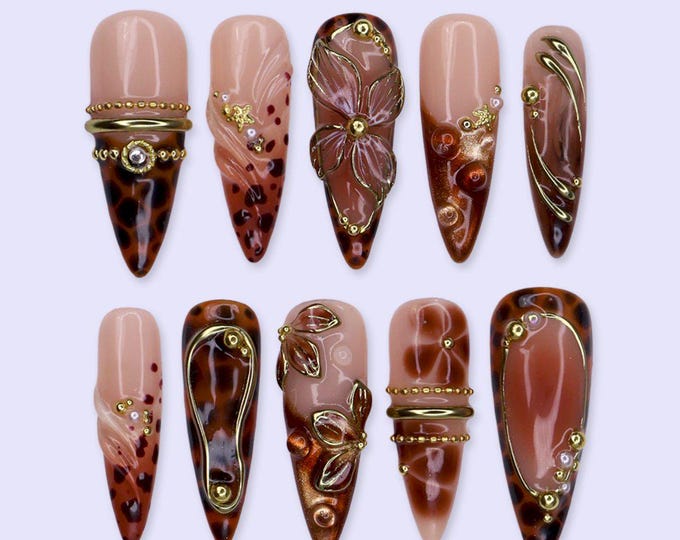 A set of ten almond-shaped press-on nails featuring a nude base with brown leopard print patterns, gold metallic accents, raised floral designs, and small pearl embellishments. The nails are arranged 