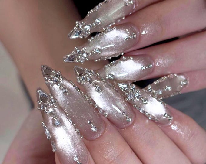Long, almond-shaped nails featuring a shimmering, pearlescent light pink polish. Each nail is intricately decorated with clusters of clear, multifaceted rhinestones and small silver beads concentrated