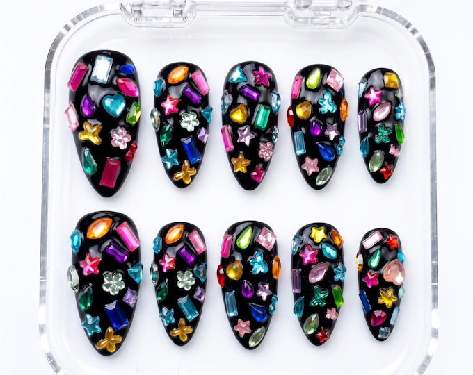 A set of ten almond-shaped press-on nails with a glossy black base, each decorated with an assortment of colorful, multi-shaped rhinestones including stars, hearts, flowers, and geometric gems, arrang