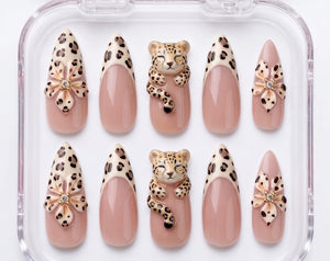 A set of ten almond-shaped press-on nails in a clear plastic case. The design features a nude base with leopard print tips, three-dimensional leopard figures, and floral accents with small embedded cr