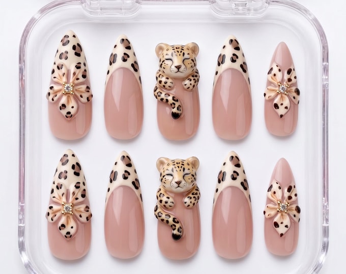 A set of ten almond-shaped press-on nails in a clear plastic case. The design features a nude base with leopard print tips, three-dimensional leopard figures, and floral accents with small embedded cr
