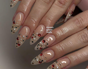 Almond-shaped nails with a glossy nude base coat, decorated with an assortment of small red, green, and black rhinestones alongside white pearls. The text moon glow nails is visible in white lettering