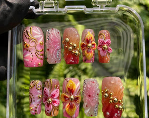 A set of ten press-on nails in a clear plastic case, featuring pink, orange, and red gradients with hand-painted floral motifs, gold metallic swirls, small gold beads, and clear rhinestones arranged i