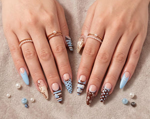 Almond-shaped nails featuring a mix of brown and light blue polish with intricate patterns including polka dots, swirls, and stripes. The nail art is accented with small white pearls and bows. Two gol