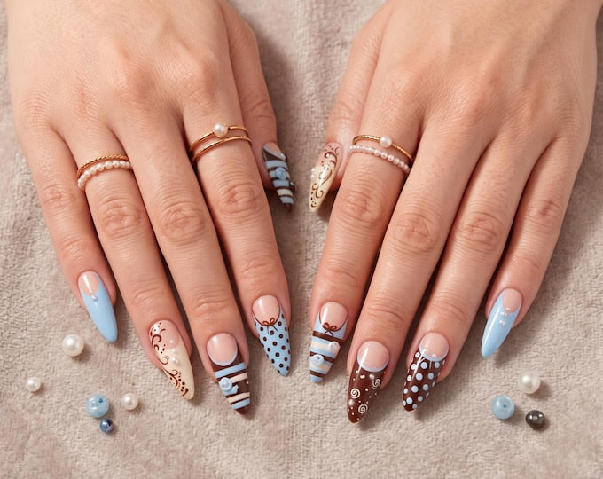 Almond-shaped nails featuring a mix of brown and light blue polish with intricate patterns including polka dots, swirls, and stripes. The nail art is accented with small white pearls and bows. Two gol