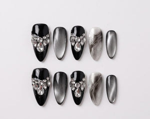 A set of ten press-on nails featuring a mix of designs. Some nails are solid black with clear rhinestone embellishments at the base, others have a grey cat-eye effect, and two feature a black and whit