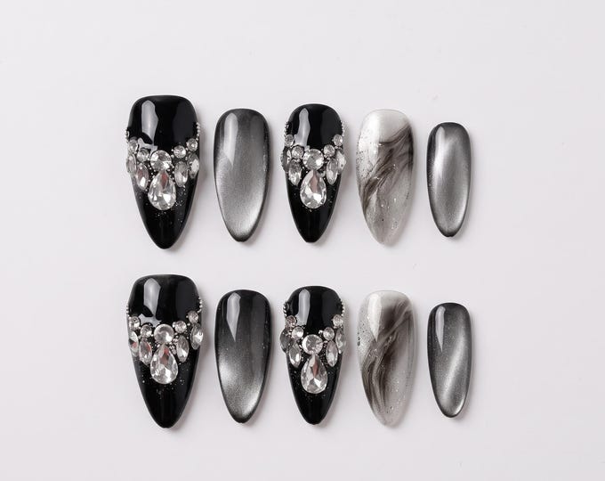 A set of ten press-on nails featuring a mix of designs. Some nails are solid black with clear rhinestone embellishments at the base, others have a grey cat-eye effect, and two feature a black and whit