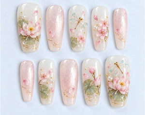A set of ten press-on nails featuring a light pink and white color palette. The designs include hand-painted pink lotus flowers, green lily pads, delicate dragonflies, and small white pearls, accented